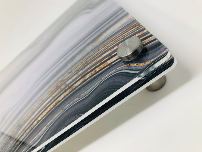 Close-up of a corner of the Raised Black Slice Challah Serving Board by Eye Candy, featuring a marbled striped pattern in gray, white, and brown tones, mounted on a cylindrical metal standoff against a white background.