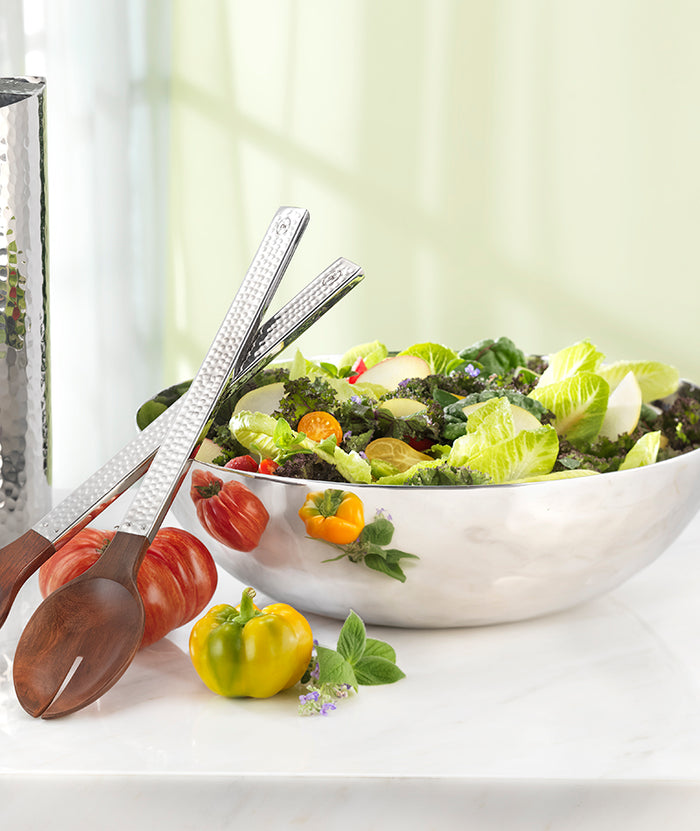 The Mary Jurek Luna Serving Bowl 12" is filled with leafy green salad and vegetables, placed on a marble surface beside wooden salad tongs, fresh tomatoes, and herbs.