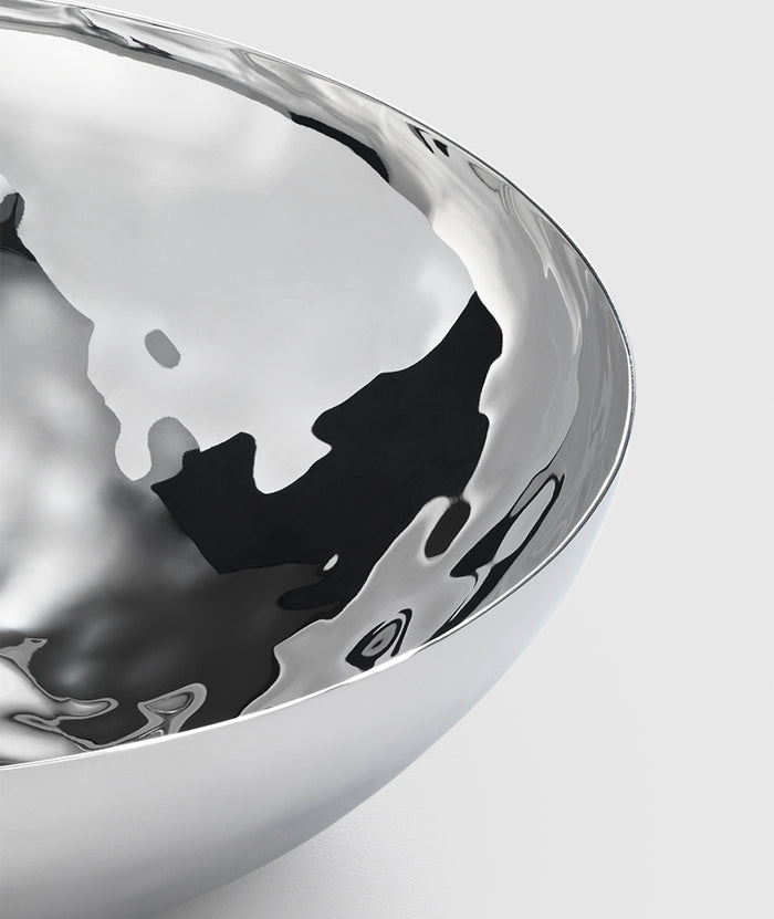 A close-up of the Mary Jurek Luna Serving Bowl 12" shows its polished, reflective stainless steel and smooth curved surfaces against a light grey background. This modern serving bowl is dishwasher safe.