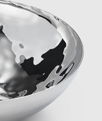 A close-up of the Mary Jurek Luna Serving Bowl 12" shows its polished, reflective stainless steel and smooth curved surfaces against a light grey background. This modern serving bowl is dishwasher safe.