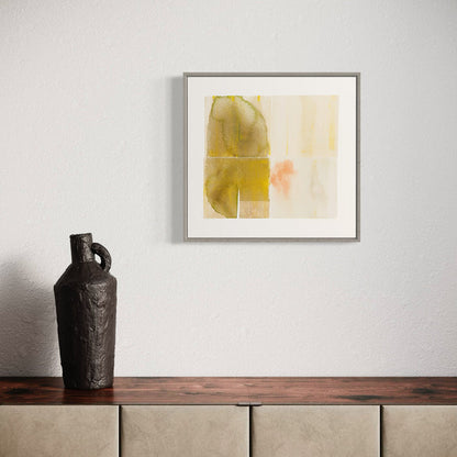 The artwork 'Placid Waters' by Ken Schiano features abstract watercolor shapes in muted olive and yellow tones, complemented by subtle peach accents on cream-white paper. The minimalist composition creates a serene, floating effect within a silver frame, measuring 21x22 inches. A contemporary watercolor that evokes tranquil, ethereal emotions.