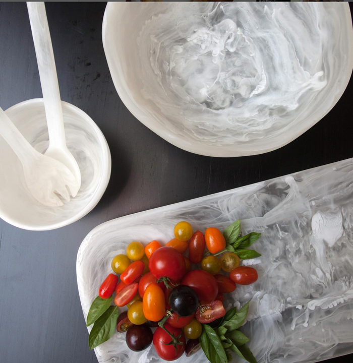 Empty bowls and a Nashi Home Swirl Classic Resin Rectangle Platter with white dressing residue, cherry tomatoes, basil, and cut tomatoes hint at a recently served salad on a dark tabletop.