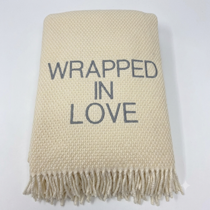 The Wrapped in Love Throw by A Soft Idea is a super soft, cream-colored woven blanket with fringed edges and "WRAPPED IN LOVE" embroidered in gray—an ideal holiday gift.