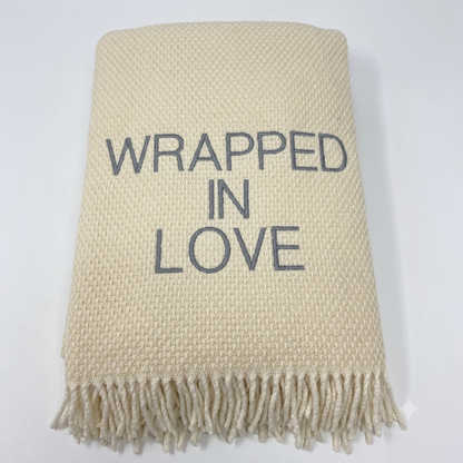 The Wrapped in Love Throw by A Soft Idea is a super soft, cream-colored woven blanket with fringed edges and "WRAPPED IN LOVE" embroidered in gray—an ideal holiday gift.