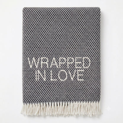A Soft Idea’s Wrapped in Love Throw is a folded dark gray woven blanket with white fringed edges. Super soft, machine washable, and “WRAPPED IN LOVE” stitched in white—perfect for gifting.